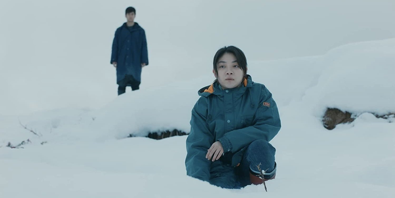 一名女子蹲在雪地里，后方站着一名男子