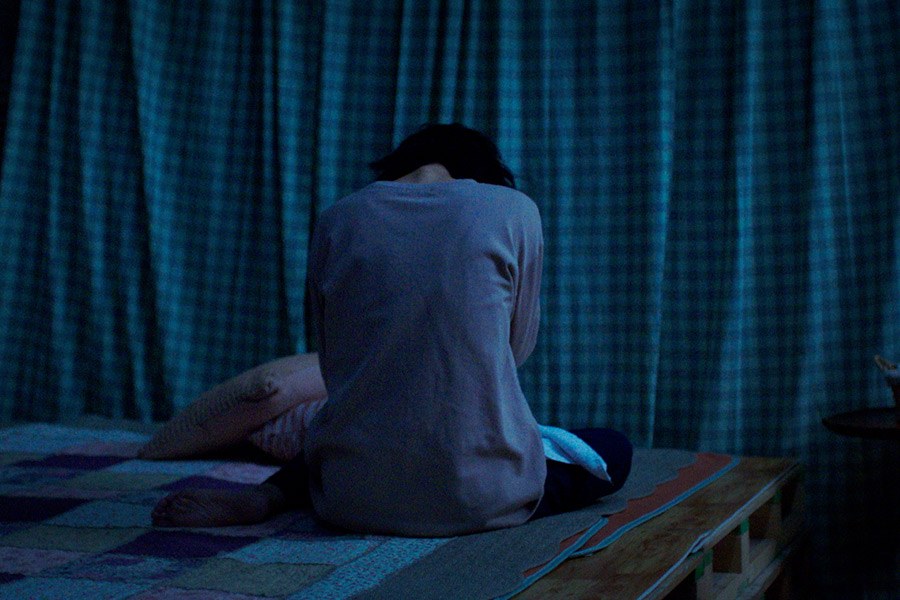 一名女子背对镜头坐在床边，身旁露出另一人的腿