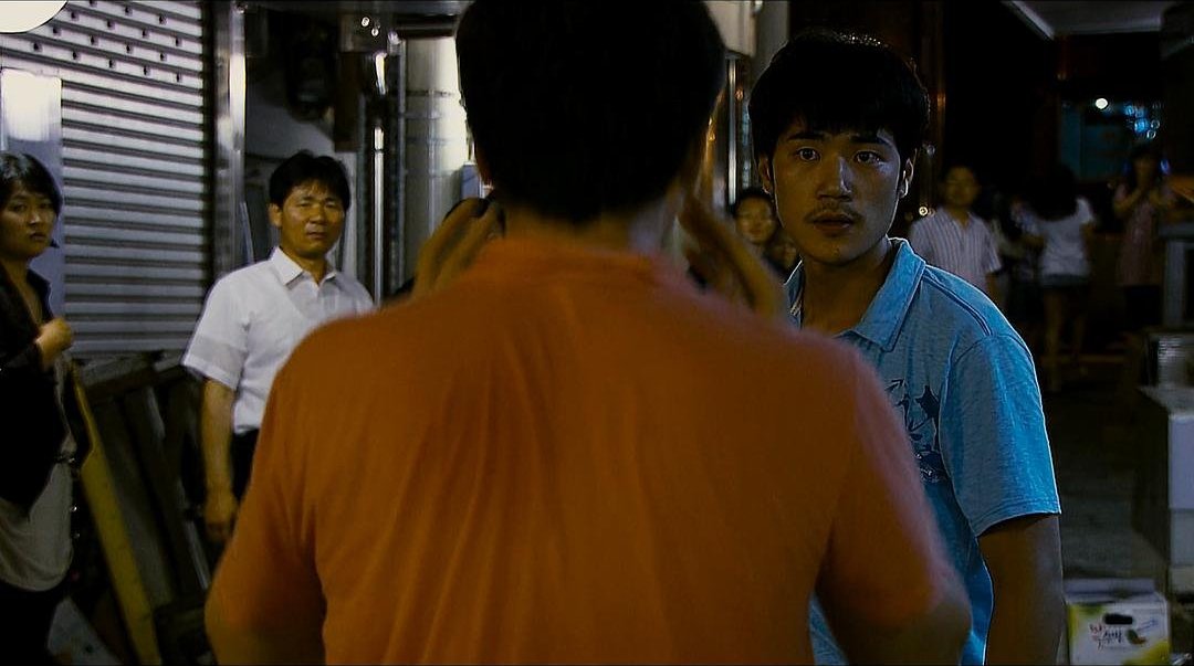 夜间街道上，一名蓝衣男子面对橙衣男子，周围有人驻足