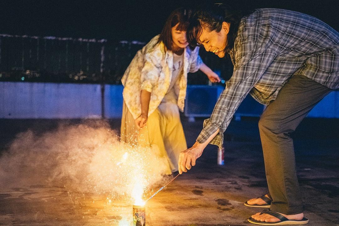 夜间屋顶上,一男一女俯身点燃地面的烟花