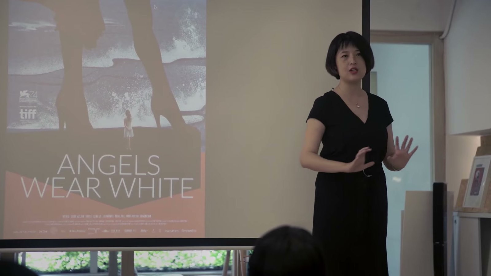 短发女性站在放映幕旁向教室里的听众讲课