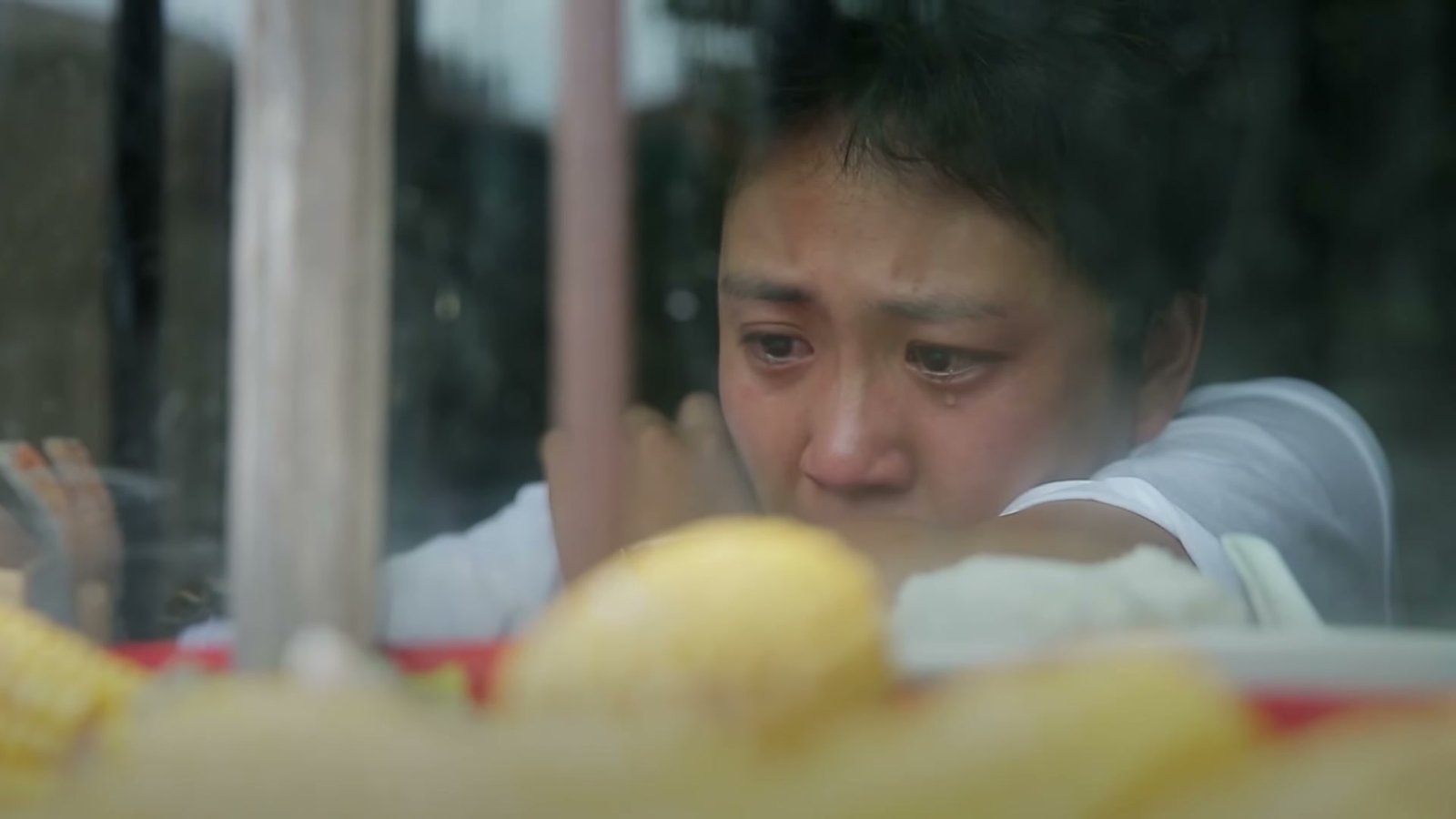 短发女性隔着窗台低头流泪，前景摆着黄色水果