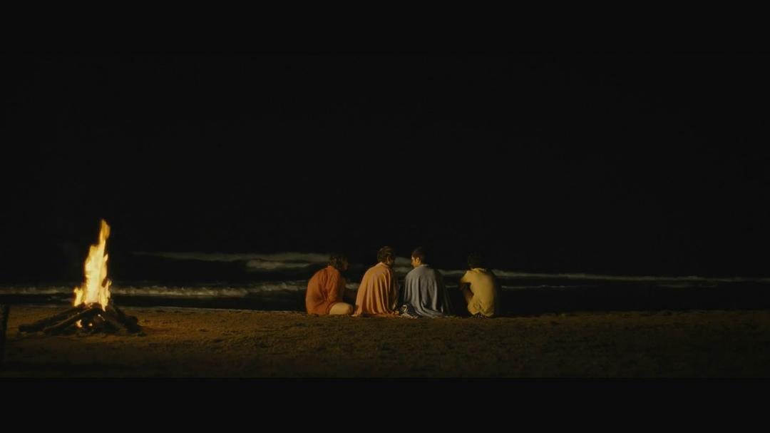 四个人背对镜头坐在夜间海滩的火堆旁