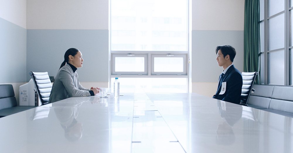 一名女面试官与一名佩戴应聘证件的男子隔着长桌交谈。