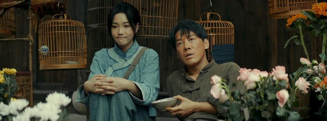 花摊前，一名女子双手交叠坐着，身旁男子捧着小碗