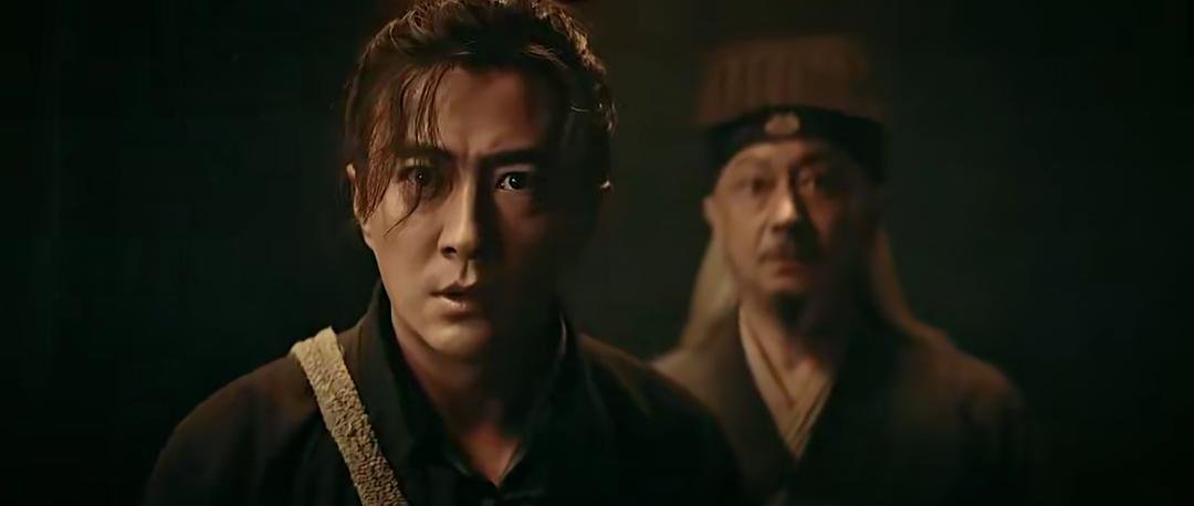 一名长发男子站在前景，另一名老者在他身后